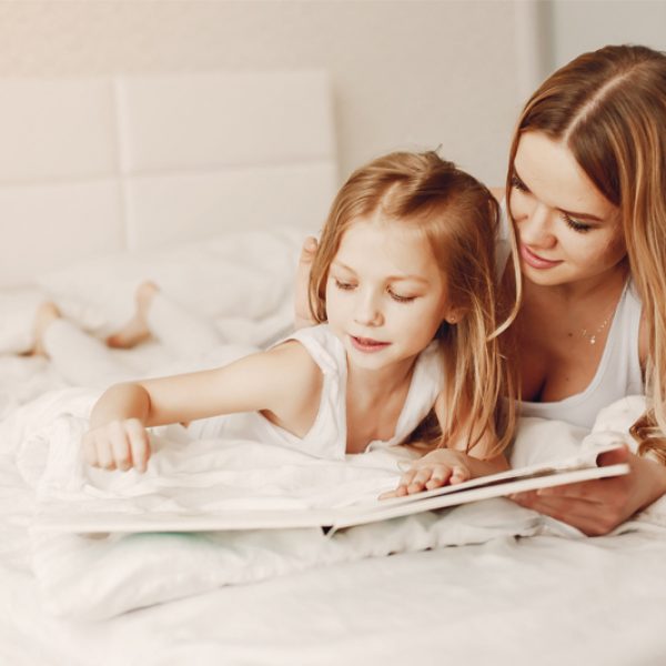 young and beautiful blonde mom reads a book in bed with her little cute daughter at home in the bedroom